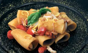Rigatoni con calamari e pomodorini pachino, guarniti con basilico e scorza di limone su piatto nero.