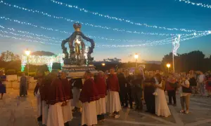 Processione serale con un carro decorato, illuminato da luci, che porta una statua. La folla osserva, mentre luci festive adornano il cielo.