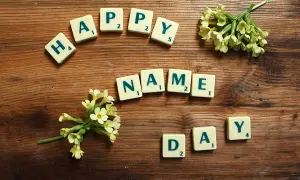 Tessere del gioco compongono "HAPPY NAME DAY" su un tavolo di legno, con fiori gialli che adornano la scena.