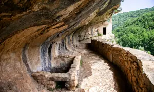 Sentiero roccioso conduce alla chiesetta di San Bartolomeo in Legio, incastonata nella roccia con vista su boschi rigogliosi.