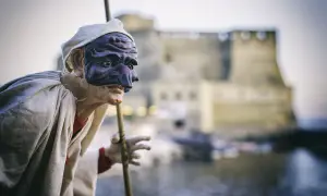 Un burattino di Pulcinella, con la sua maschera iconica, si staglia contro un castello sfocato. Una pulcinella storia che evoca tradizioni napoletane.