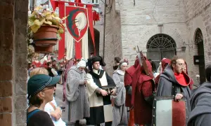 Persone in costume medievale sfilano in una strada acciottolata, con stendardi rossi e bianchi che sventolano. La scena evoca il **calendimaggio significato**, una festa di primavera.