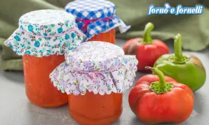 Barattoli di conserva di peperoni napoletana con tappi decorati, circondati da peperoni freschi rossi e verdi.