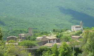 Il gigante di acciaio veglia su un borgo antico, tra torri e case di pietra, sotto un monte verdeggiante.