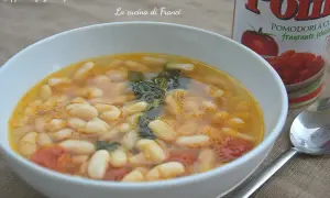 Zuppa di fagioli spollichini fumante in una ciotola bianca, con pomodori e verdure. Accanto, una lattina di pomodori e un cucchiaio.
