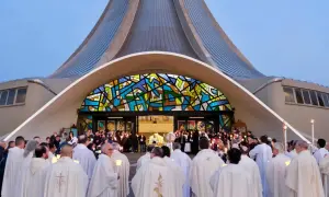 Un gruppo di sacerdoti, con candele accese, si riunisce davanti a una chiesa moderna con una vetrata colorata. La luce delle candele illumina i loro volti, quasi a riflettere il volto di Gesù.