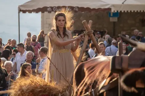 Donna in abito dorato, con spighe di grano che simboleggiano abbondanza e fertilità, durante una festa popolare.
