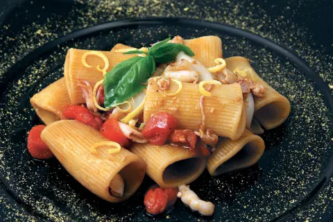 Rigatoni con calamari e pomodorini pachino, guarniti con basilico e scorza di limone su piatto nero.