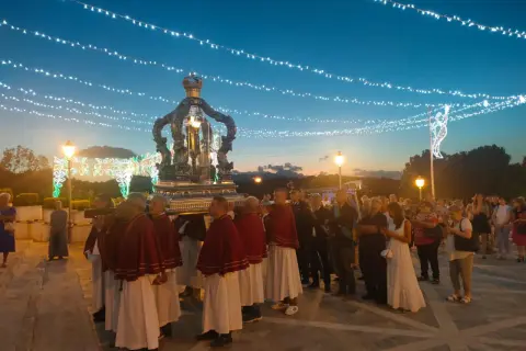 Processione serale con un carro decorato, illuminato da luci, che porta una statua. La folla osserva, mentre luci festive adornano il cielo.