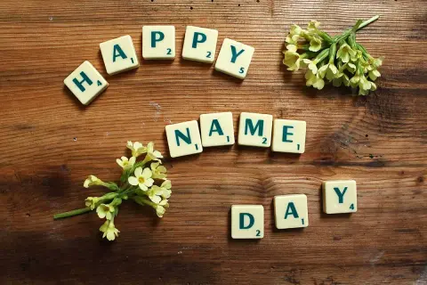 Tessere del gioco compongono "HAPPY NAME DAY" su un tavolo di legno, con fiori gialli che adornano la scena.