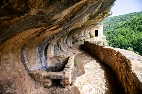 Sentiero roccioso conduce alla chiesetta di San Bartolomeo in Legio, incastonata nella roccia con vista su boschi rigogliosi.