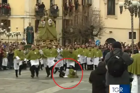 Durante una processione, un uomo cade a terra, quasi come una **madonna che scappa**, mentre altri partecipano con vesti verdi e bianche.