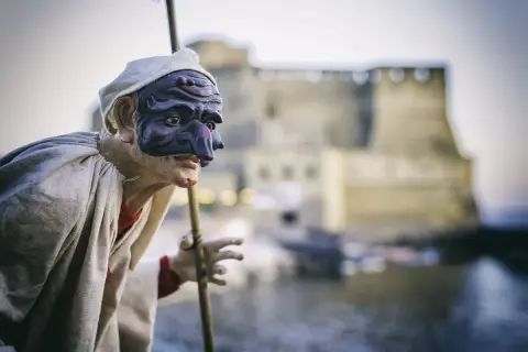 Un burattino di Pulcinella, con la sua maschera iconica, si staglia contro un castello sfocato. Una pulcinella storia che evoca tradizioni napoletane.