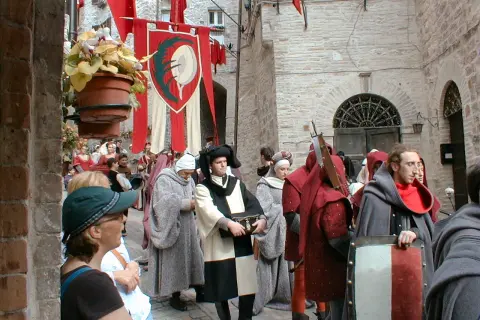 Persone in costume medievale sfilano in una strada acciottolata, con stendardi rossi e bianchi che sventolano. La scena evoca il **calendimaggio significato**, una festa di primavera.