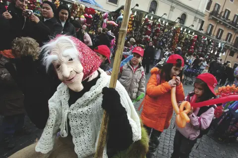 Una maschera della Befana saluta la folla, mentre i bambini giocano con palloncini gonfiabili. Si festeggia la Befana!