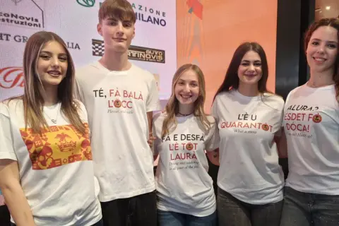 Cinque giovani sorridono, indossando magliette con scritte in dialetto. Una dice "Té, fà balà l'oeucc", che significa "Ti faccio l'occhiolino", un invito a prendersi cura di sé.