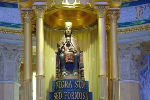 Statua dorata di una Maria nera con Gesù Bambino, su un piedistallo con iscrizione latina.