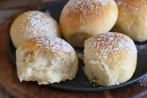 Cinque soffici panini spolverati di zucchero a velo, uno è stato morso rivelando la sua brioche rustica morbidissima.