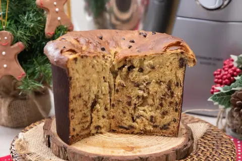 Un soffice panettone natalizio con gocce di cioccolato, tagliato a metà, su un tagliere di legno. Decorazioni natalizie sullo sfondo.