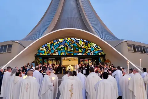 Un gruppo di sacerdoti, con candele accese, si riunisce davanti a una chiesa moderna con una vetrata colorata. La luce delle candele illumina i loro volti, quasi a riflettere il volto di Gesù.