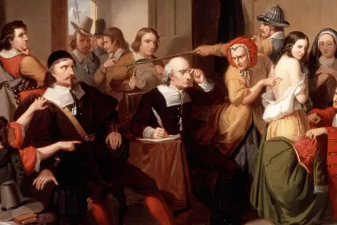 Un gruppo di persone esamina una giovane donna, cercando segni fisici delle streghe. Un uomo scrive, mentre altri osservano con sospetto.