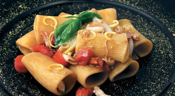Rigatoni con calamari e pomodorini pachino, guarniti con basilico e scorza di limone su piatto nero.