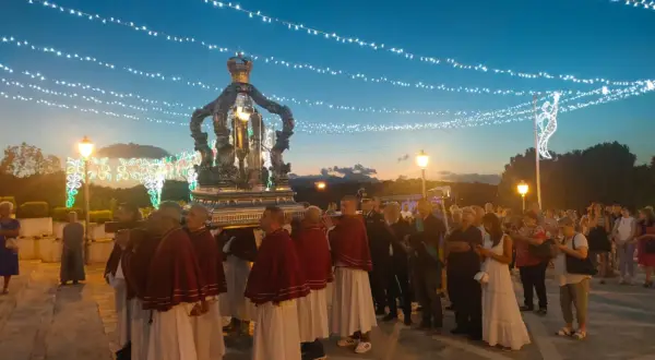 Processione serale con un carro decorato, illuminato da luci, che porta una statua. La folla osserva, mentre luci festive adornano il cielo.