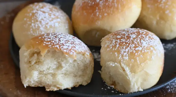 Cinque soffici panini spolverati di zucchero a velo, uno è stato morso rivelando la sua brioche rustica morbidissima.