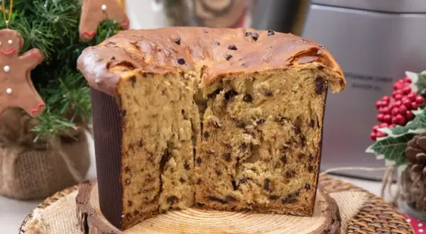 Un soffice panettone natalizio con gocce di cioccolato, tagliato a metà, su un tagliere di legno. Decorazioni natalizie sullo sfondo.