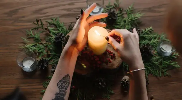 Accensione di una candela, simbolo della ruota dell'anno. Mani con tatuaggi e braccialetto, circondate da pigne e rami.