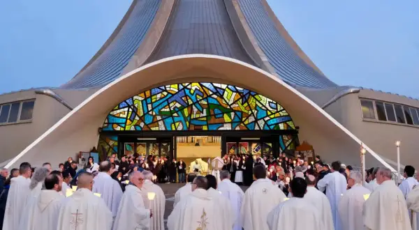 Un gruppo di sacerdoti, con candele accese, si riunisce davanti a una chiesa moderna con una vetrata colorata. La luce delle candele illumina i loro volti, quasi a riflettere il volto di Gesù.