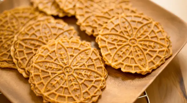 Pizzelle abruzzesi dorate e decorate con un motivo a fiore, disposte su un piatto di legno.