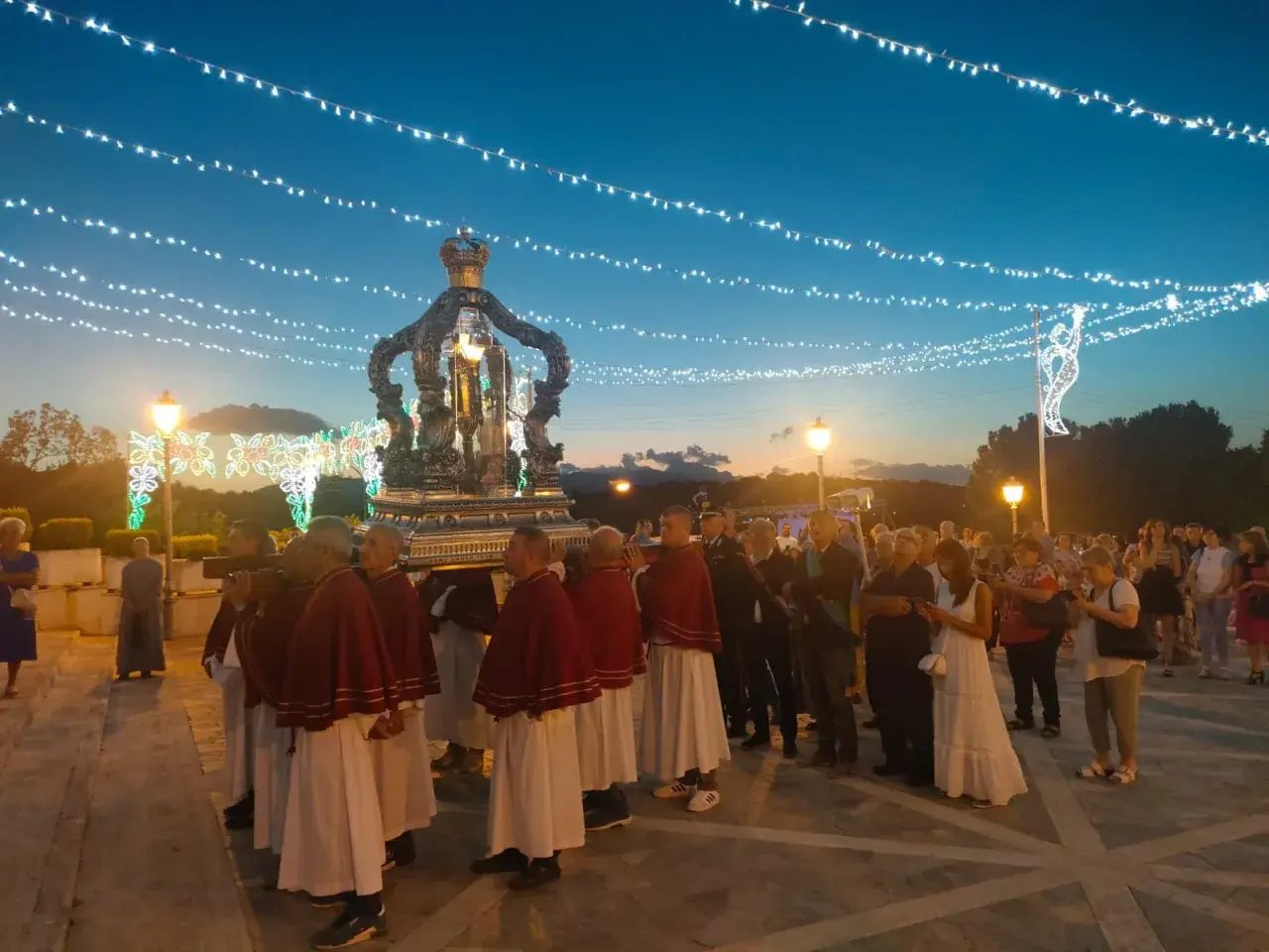 Processione serale con un carro decorato, illuminato da luci, che porta una statua. La folla osserva, mentre luci festive adornano il cielo.
