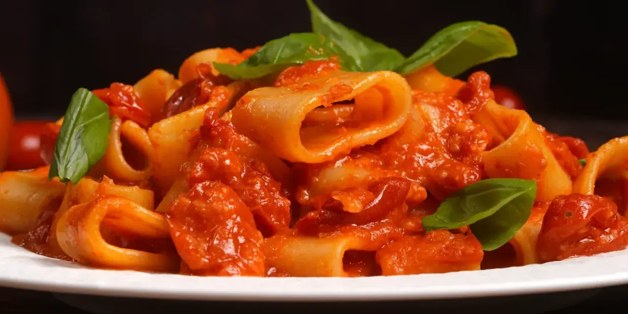 Un piatto di rigatoni allo scarpariello, conditi con un sugo rosso ricco e guarniti con foglie di basilico fresco.
