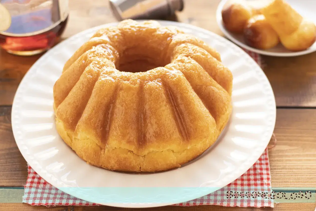 Un soffice babà, pronto per essere inzuppato, evoca la **babà ricetta originale**. Accanto, un bicchierino di rum e altri babà in miniatura.