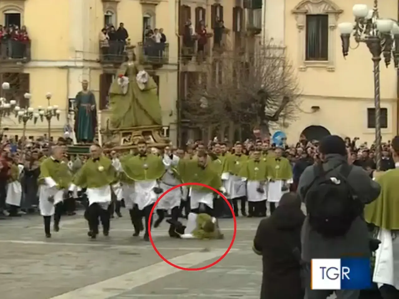 Durante una processione, un uomo cade a terra, quasi come una **madonna che scappa**, mentre altri partecipano con vesti verdi e bianche.