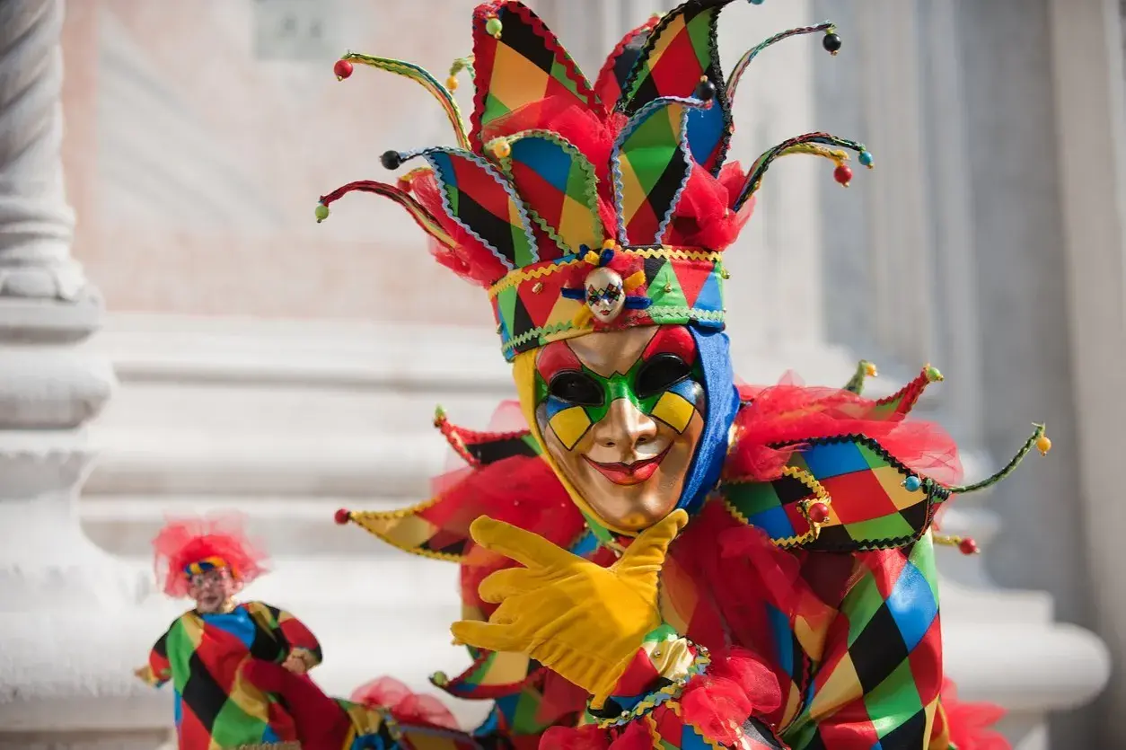 Un personaggio con un elaborato tipo di maschera di carnevale, colorato e festoso, con un cappello a punta e guanti gialli.