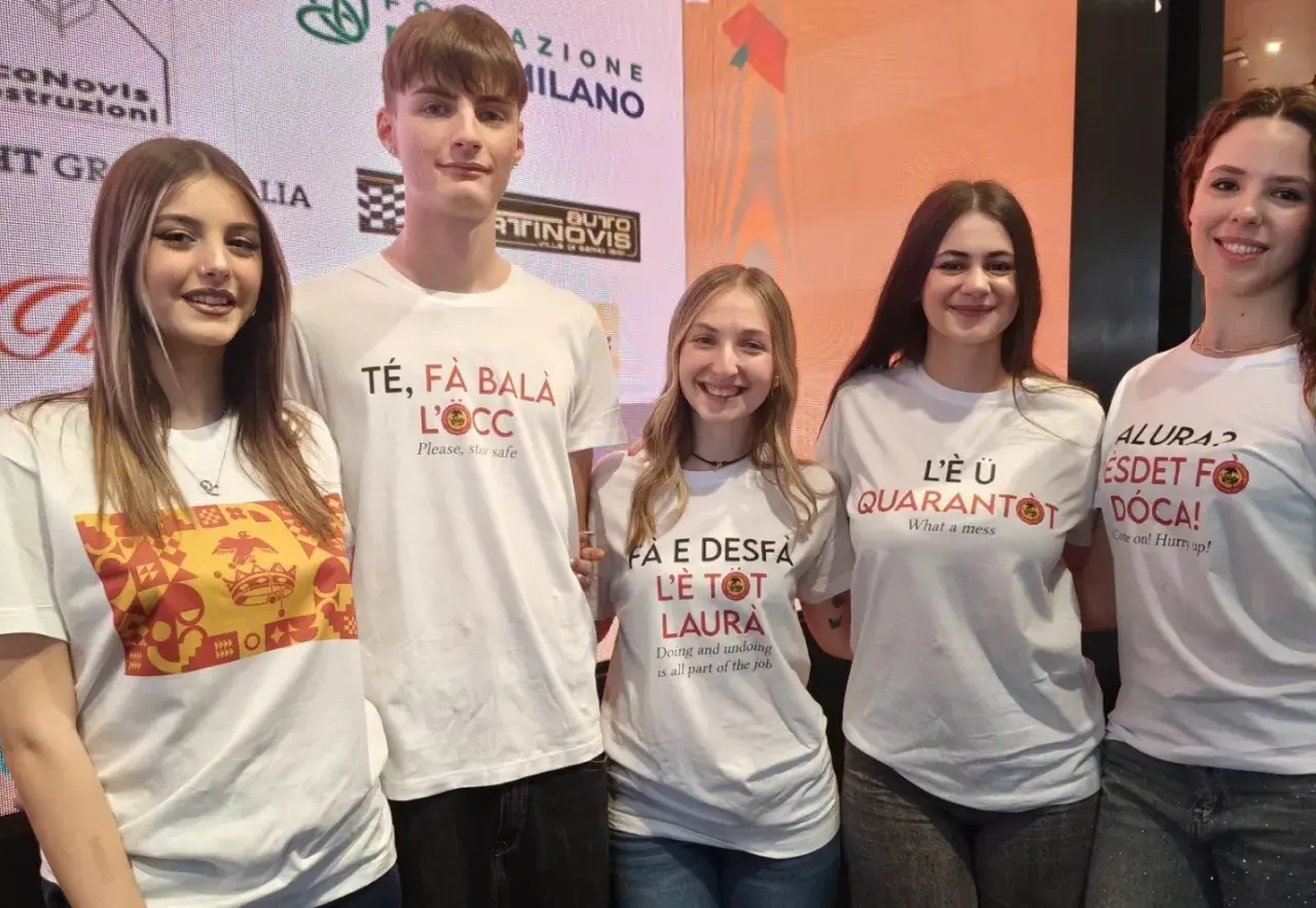 Cinque giovani sorridono, indossando magliette con scritte in dialetto. Una dice "Té, fà balà l'oeucc", che significa "Ti faccio l'occhiolino", un invito a prendersi cura di sé.