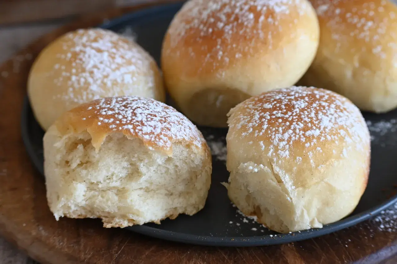 Cinque soffici panini spolverati di zucchero a velo, uno è stato morso rivelando la sua brioche rustica morbidissima.