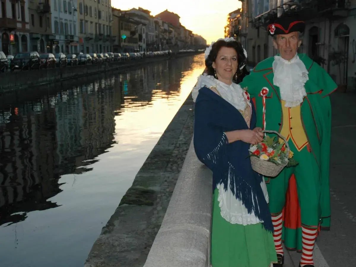 Coppia in costume tradizionale, una maschera milanese, passeggia lungo un naviglio al tramonto.