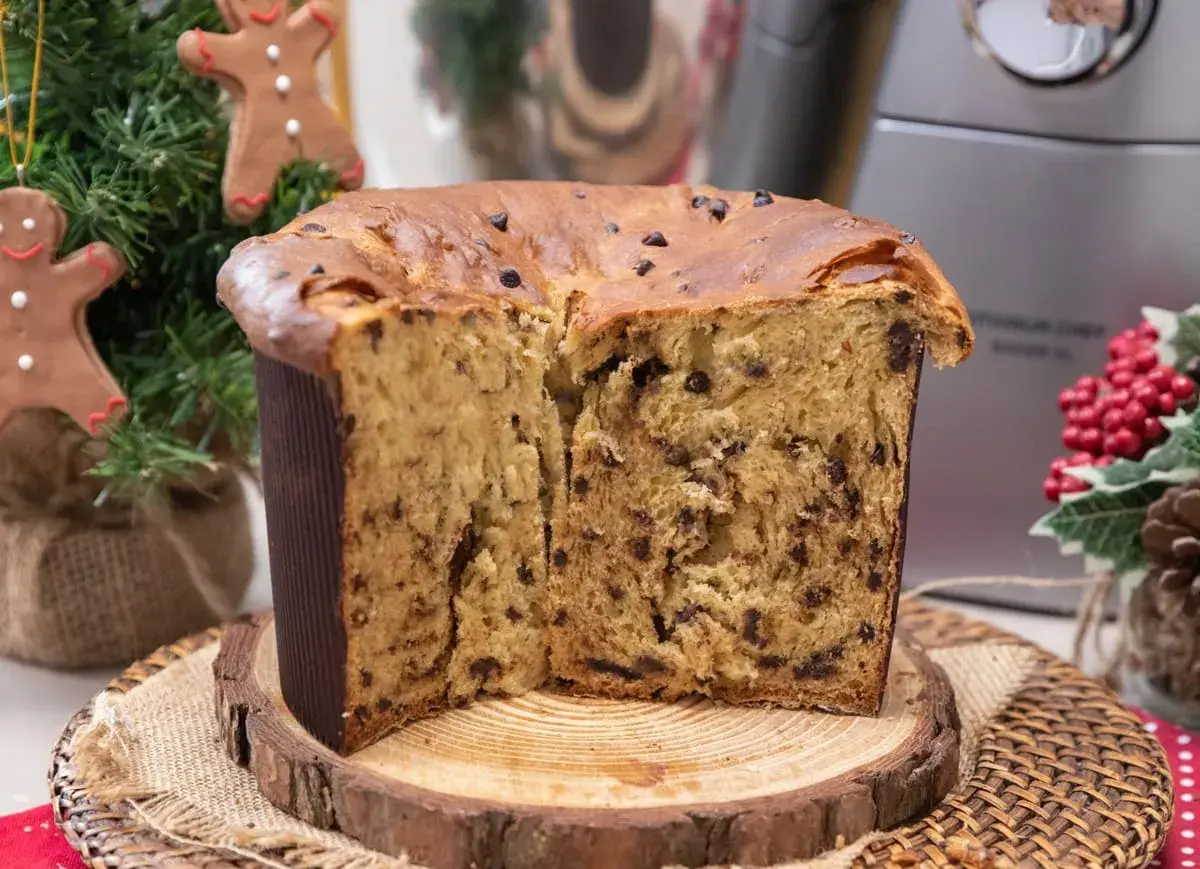 Un soffice panettone natalizio con gocce di cioccolato, tagliato a metà, su un tagliere di legno. Decorazioni natalizie sullo sfondo.