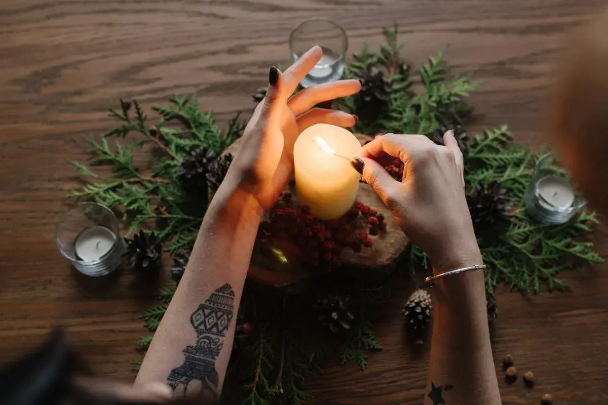 Accensione di una candela, simbolo della ruota dell'anno. Mani con tatuaggi e braccialetto, circondate da pigne e rami.