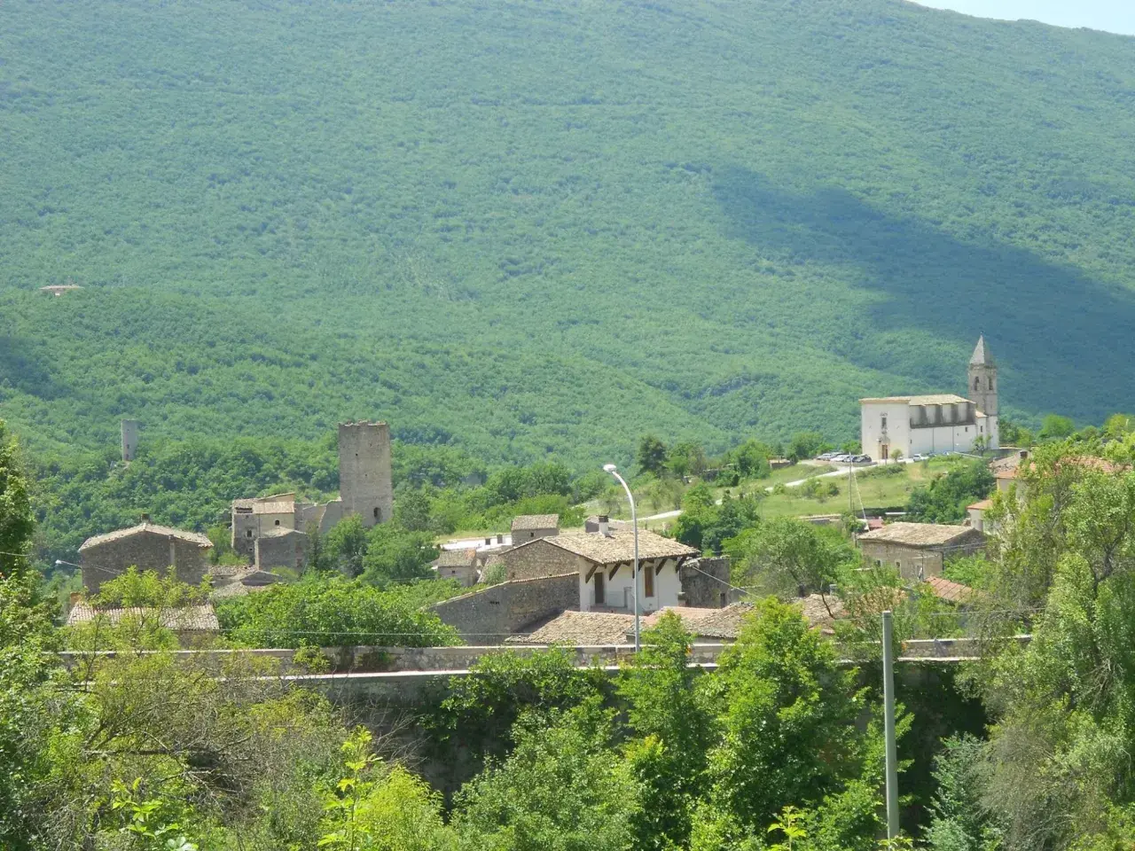 Il gigante di acciaio veglia su un borgo antico, tra torri e case di pietra, sotto un monte verdeggiante.