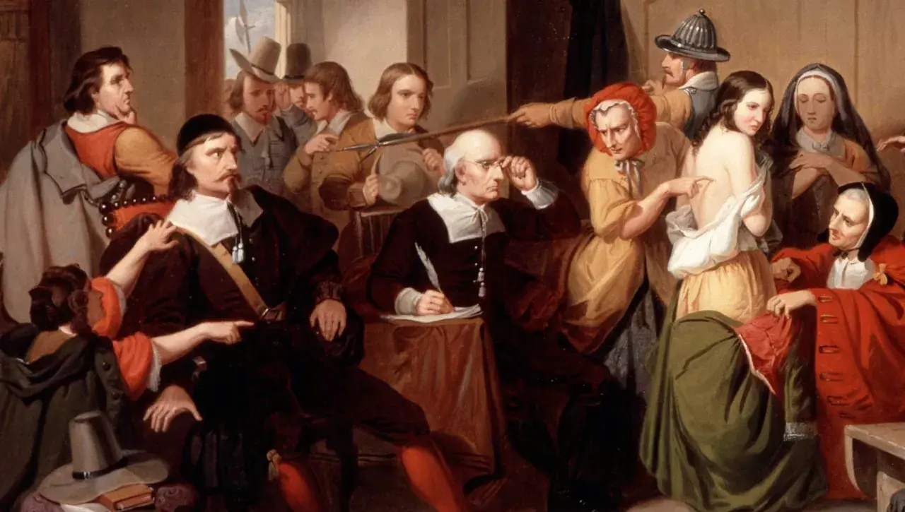 Un gruppo di persone esamina una giovane donna, cercando segni fisici delle streghe. Un uomo scrive, mentre altri osservano con sospetto.