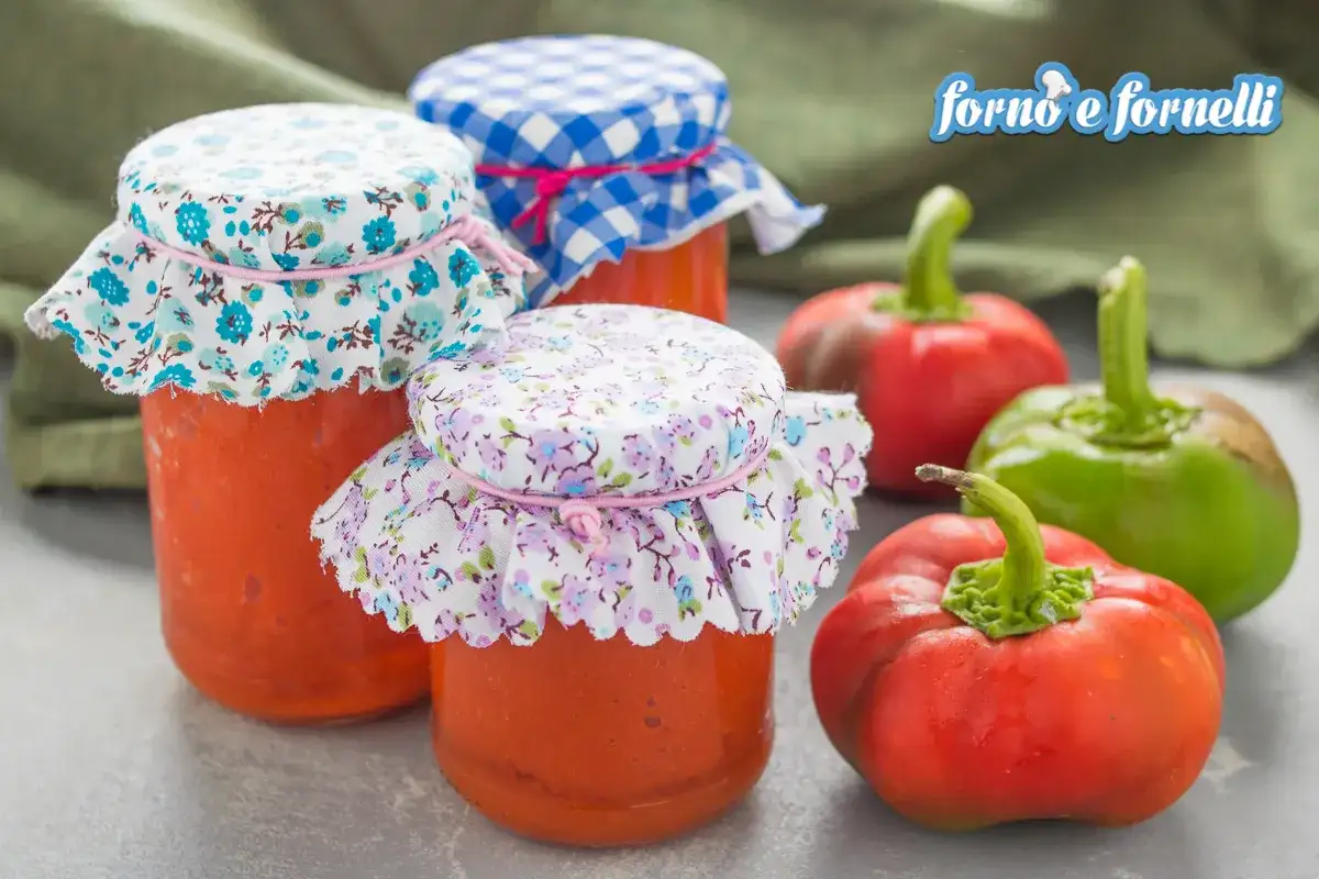 Barattoli di conserva di peperoni napoletana con tappi decorati, circondati da peperoni freschi rossi e verdi.