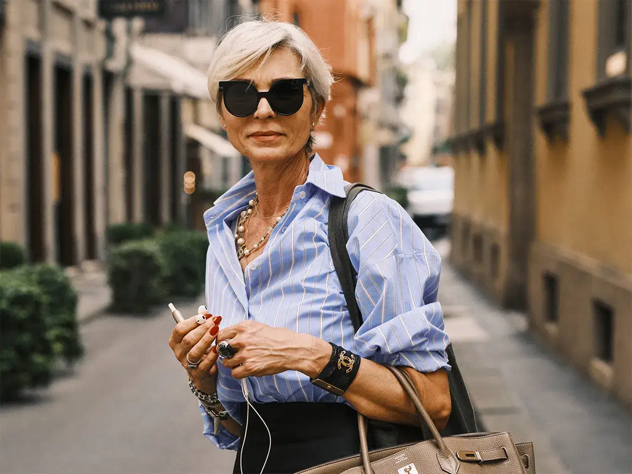 Una sciura milanese con occhiali da sole, camicia a righe e gioielli, in una strada acciottolata.