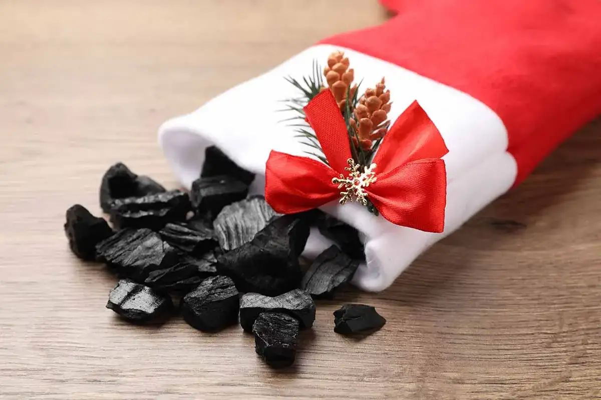 Un calzino rosso e bianco decorato con un fiocco rosso e pigne, rovesciato su un tavolo di legno, con pezzi di carbone che fuoriescono. La Befana porta via le feste con un sacco di carbone.