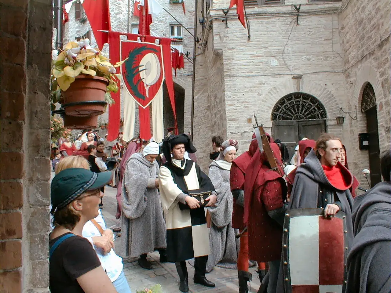 Persone in costume medievale sfilano in una strada acciottolata, con stendardi che evocano il calendimaggio significato.