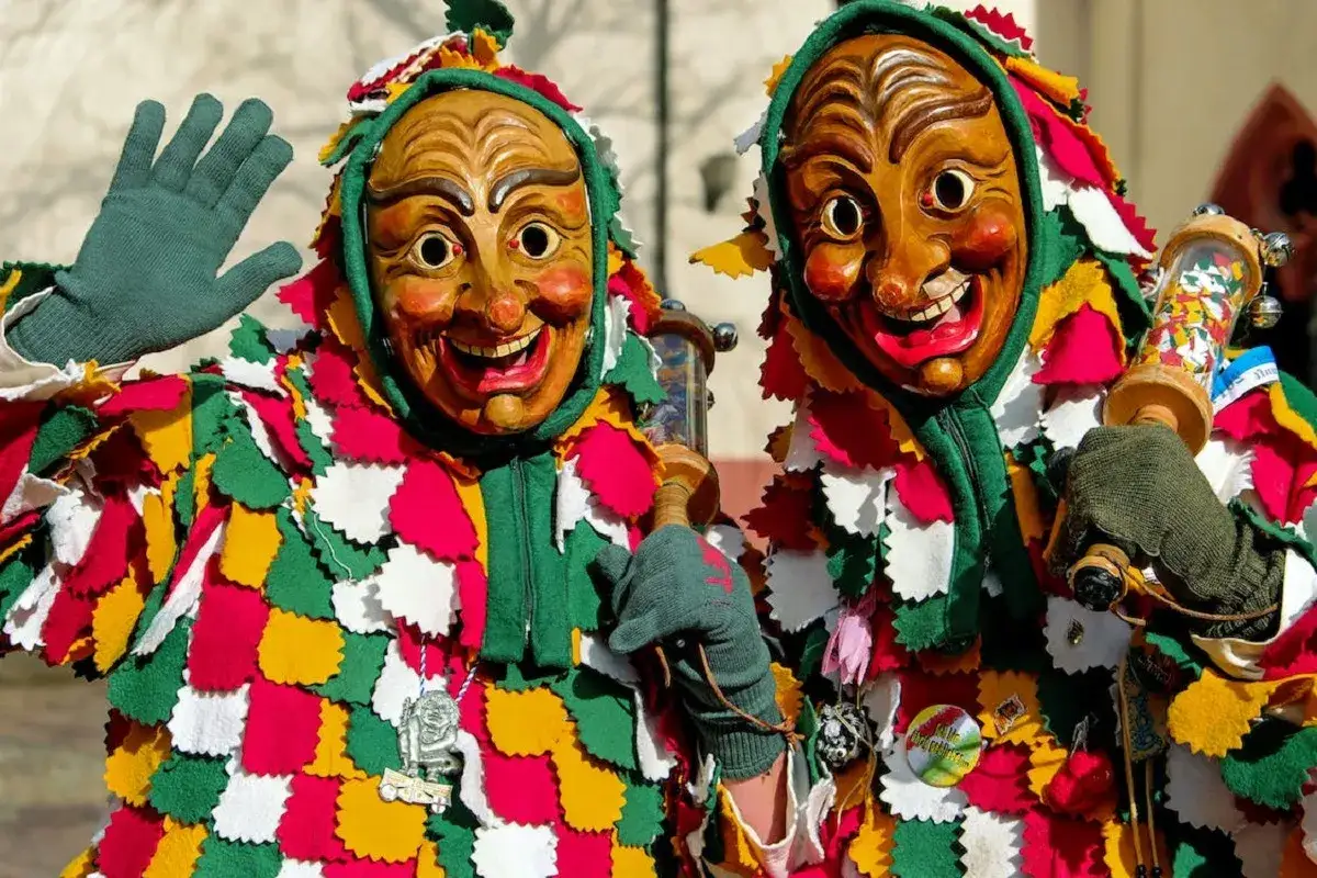 Due maschere di legno sorridono, vestite con colorati costumi che ricordano una festa pagana di carnevale. Una saluta con la mano.