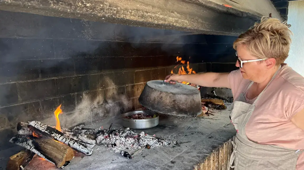 Donna prepara patate al coppo in un forno a legna. La pentola di metallo è sollevata, rivelando il cibo.